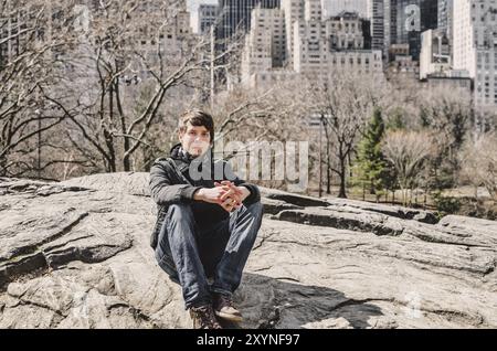 Jeune homme adulte aux cheveux bruns et à la barbe regardant la caméra tout en étant assis sur un rocher dans le parc central, horizon de manhattan New york dans l'arrière-plan Banque D'Images