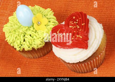 Deux cupcakes de vacances de fantaisie sur un fond orange. Un cupcake de Saint-Valentin avec un coeur rouge crème au beurre sur la droite et une décoration cupcake de Pâques Banque D'Images