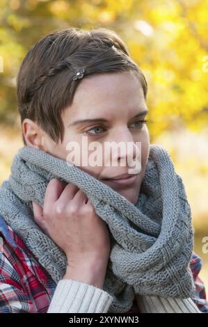 Jolie femme pensive aux cheveux courts se blottissant dans une écharpe d'hiver tricotée chaude. Feuillage d'automne coloré en arrière-plan Banque D'Images