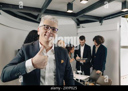 Homme d'affaires mature souriant montrant le pouce vers le haut geste tout en se tenant debout dans un bureau moderne avec divers collègues travaillant sur le projet Banque D'Images