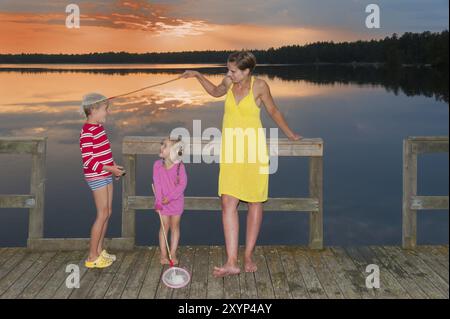 Jeune mère debout avec ses enfants sur une jetée au bord d'un lac à Smaland, en Suède, profitant de la belle soirée d'été. Elle joue et trompe aro Banque D'Images