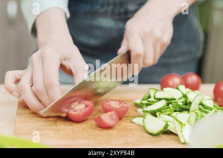 Gros plan les mains féminines délicates coupent un grand couteau avec des tomates sur un quart sur une planche de bois à la maison. Cuisine domestique. Alimentation saine. Le concept de ve Banque D'Images