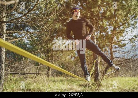 Un homme, vieilli avec une barbe et portant des lunettes de soleil, se balance sur une slackline en plein air entre deux arbres au coucher du soleil sur fond bleu ciel Banque D'Images