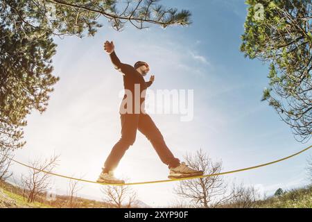 Un homme, vieilli avec une barbe et portant des lunettes de soleil, se balance sur une slackline en plein air entre deux arbres au coucher du soleil sur fond bleu ciel Banque D'Images