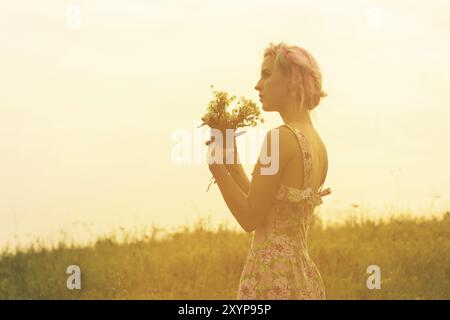 Jeune femme vêtue avec bouquet de fleurs entre les mains au coucher du soleil dans le champ. Image teintée et chaude Banque D'Images