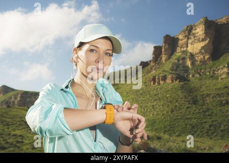 Portrait d'une jeune fille de fitness souriante dans une casquette et des écouteurs vérifiant son horloge intelligente tout en étant assise à l'extérieur sur un fond de rochers. Lookin Banque D'Images