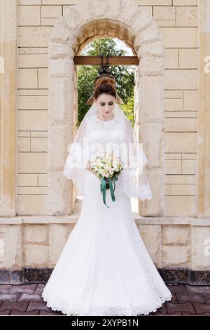 Une belle mariée dans une robe de mariée et un bouquet de mariage dans ses mains se tient à l'extérieur contre une arche de pierre avec une cloche. Le concept de happi de mariage Banque D'Images