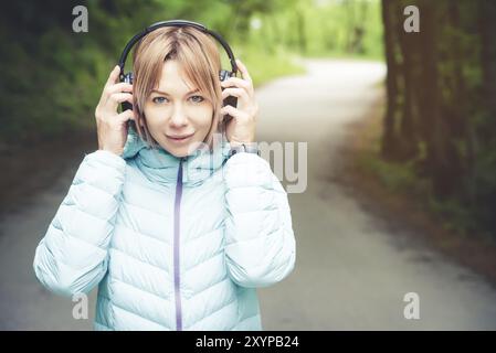 Portrait d'une fille de sport attrayante blonde dans une veste de descente légère habillant casque bluetooth avec de la musique ou les sons de la nature tout en étant sur Banque D'Images