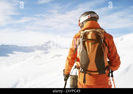 Un skieur dans une combinaison orange avec un sac à dos sur le dos portant un casque et avec des bâtons de ski dans ses mains se tient debout sur un précipice devant un sno Banque D'Images