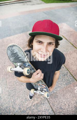 Un jeune patineur aux cheveux longs portant un T-shirt et une casquette pleine longueur se tient debout avec une planche à roulettes sur le fond du paysage urbain Banque D'Images