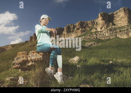 Portrait d'une jeune fille de fitness souriante dans une casquette et des écouteurs vérifiant son horloge intelligente assise sur un rocher à l'extérieur sur un fond de rochers Banque D'Images