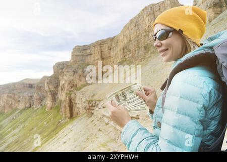 Un voyageur dans un chapeau et des lunettes de soleil tient une centaine de billets de dollars dans les mains d'un ventilateur sur fond de rochers sur la nature. Frais de déplacement Banque D'Images