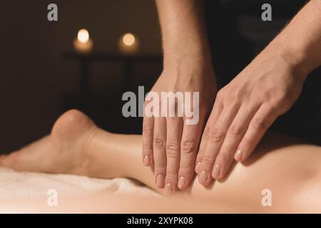 Gros plan des mains masculines faisant massage des mollets des jambes féminines dans une pièce sombre avec des bougies en arrière-plan. Cosmétologie et soins spa Banque D'Images