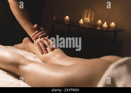 Gros plan des mains masculines faisant massage des mollets des jambes féminines dans une pièce sombre avec des bougies en arrière-plan. Cosmétologie et soins spa Banque D'Images