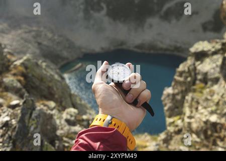 La main d'un homme tient une boussole magnétique de poche pour la navigation sur la toile de fond d'une pente rocheuse et d'un lac de montagne. Le concept de trouver un moyen. G Banque D'Images