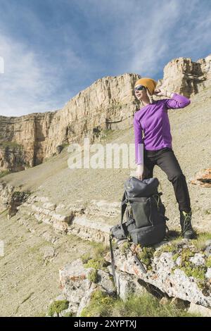 Voyageuse fille souriante dans un chapeau jaune et une paire de lunettes de soleil se tient au pied de rochers épiques avec un sac à dos à côté et regarde loin Banque D'Images