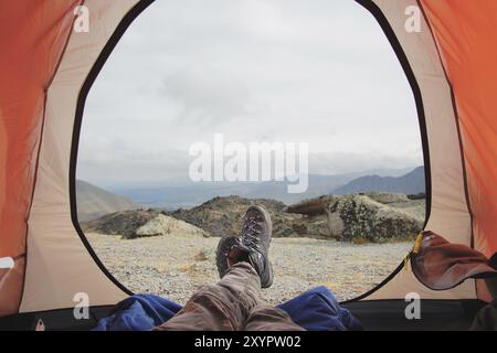 Jambes d'un homme reposant dans la tente de voyage dans des bottes pour le suivi de montagne sur fond de montagnes et de vallées avec des nuages bruyants bottes de randonnée en th Banque D'Images