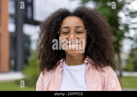 Étudiante afro-américaine confiante avec des cheveux bouclés et des lunettes souriant chaleureusement à l'extérieur. Capture l'essence de la jeunesse et de l'éducation, en soulignant l'attitude positive et le comportement amical Banque D'Images