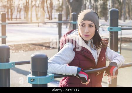 Portrait d'une athlète fille dans un chapeau avec des gants et un gilet chaud à côté d'une aire de jeux d'entraînement en plein air en hiver par une journée ensoleillée Banque D'Images