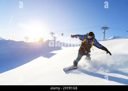 Un snowboarder dans un masque de ski et un sac à dos roule sur une pente enneigée laissant derrière lui une poudre de neige contre le ciel bleu et le soleil couchant. Banque D'Images