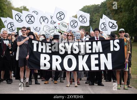 Windsor, Royaume-Uni. 30 août 2024. Extinction Rebellion a organisé une manifestation sur la longue marche avec pour toile de fond le château de Windsor à Windsor, Berkshire ce soir. Les manifestants climatiques de XR portaient des costumes d'affaires et s'habillaient en barons du pétrole, en magnats des médias et en financiers sans visage. Les Oil Slicks, habillés de costumes noirs avec de la peinture de visage noir et blanc, ont réalisé une action théâtrale chorégraphiée dans le but de mettre en évidence le récit XR pour la journée, que le système est brisé. Les rebelles XR campent à Home Park en face du château de Windsor aujourd'hui et pendant le week-end dans le cadre de leur surclassement de trois jours Banque D'Images