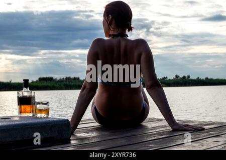 Une femme profite du coucher de soleil au bord de l'eau sur un quai en bois avec une bouteille et un verre de spiritueux Banque D'Images