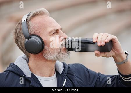 Gros plan portrait de l'homme aux cheveux blancs senior dans des écouteurs tenant une bouteille en plastique d'eau potable tout en se reposant après le jogging dans le parc de la ville Banque D'Images