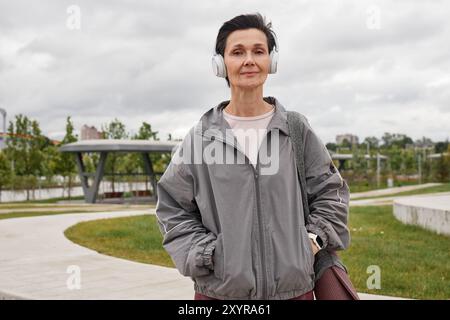 Portrait moyen d'une femme âgée souriante dans des écouteurs portant un coupe-vent gris et regardant la caméra pendant l'entraînement quotidien dans un parc venteux avec ciel couvert, espace copie Banque D'Images