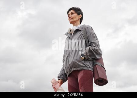 Plein plan moyen d'une femme âgée souriante avec des écouteurs portant des vêtements de sport tenant une bouteille d'eau tout en se reposant après l'entraînement matinal dans le parc de la ville venteux contre le ciel couvert gris, espace de copie Banque D'Images