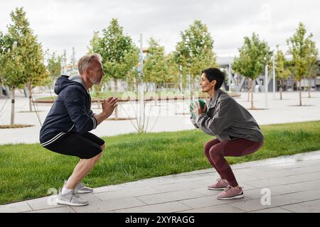 Vue latérale de la femme et de l'homme souriant senior en vêtements de sport faisant squat sauts de balle passant Banque D'Images