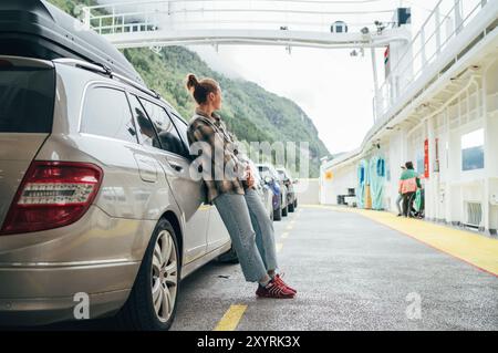 Femme s'appuie contre sa voiture sur le ferry norvégien entouré par d'autres véhicules avec des montagnes vertes brumeuses en arrière-plan. Scène capture l'essence de relaxe Banque D'Images