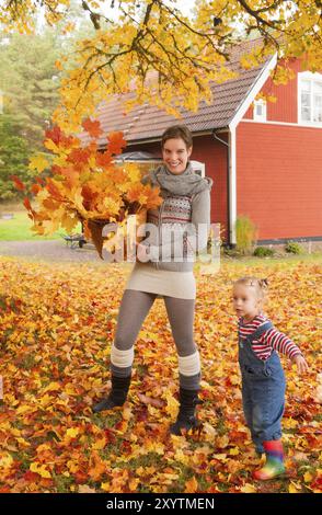 La jeune mère et sa petite fille ramassent les feuilles d'automne ensemble dans un panier et profitent de l'automne doré. En arrière-plan un HO en bois rouge typique Banque D'Images