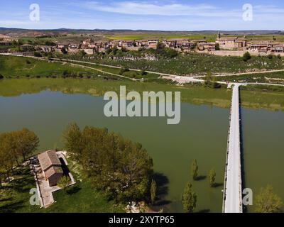 Une vue aérienne d'une rivière avec un pont menant à un village avec de nombreuses maisons, entouré de végétation sous un ciel bleu, vue aérienne, Maderuelo, Rio Banque D'Images