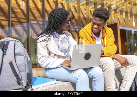 Jeunes amis d'université afro-américains utilisant un ordinateur portable assis à l'extérieur du campus dans une journée ensoleillée Banque D'Images