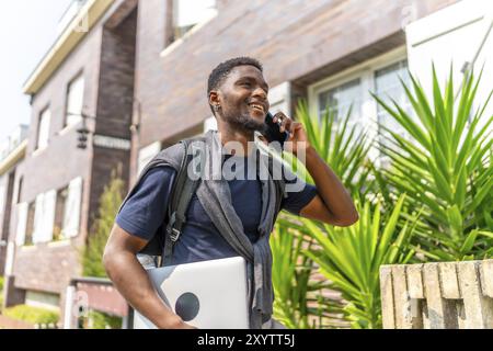 Homme africain transportant un ordinateur portable et utilisant un mobile dans la rue marchant le long d'un quartier résidentiel Banque D'Images