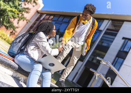 Étudiante africaine saluant un ami tout en utilisant un ordinateur portable assis dans le campus à l'extérieur Banque D'Images