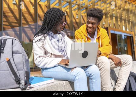 Deux jeunes étudiants africains, hommes et femmes, souriants tout en utilisant un ordinateur portable assis à l'extérieur du campus universitaire Banque D'Images