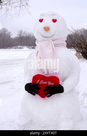 Bonhomme de neige souriant avec des yeux en forme de cœur tenant un cœur rouge avec I Love You écrit dessus dans un paysage hivernal couvert de neige. Saint Valentin romantique Conce Banque D'Images