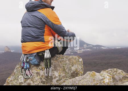 Hipster, un grimpeur en veste duvet et un bonnet tricoté s'assoit et repose sur un rocher avec une vue sur le ponamram automnal des montagnes voisines an Banque D'Images