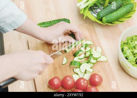 Gros plan des mains femelles coupées en concombres frais coupés sur une planche à découper en bois à côté des tomates roses. Le concept de cuisine végétarienne maison et Banque D'Images