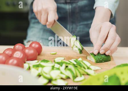 Gros plan des mains femelles coupées en concombres frais coupés sur une planche à découper en bois à côté des tomates roses. Le concept de cuisine végétarienne maison et Banque D'Images