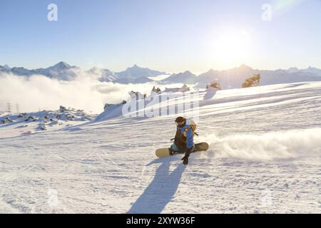 Un snowboarder dans un masque de ski et un sac à dos roule sur une pente enneigée laissant derrière lui une poudre de neige contre le ciel bleu et le soleil couchant. Banque D'Images