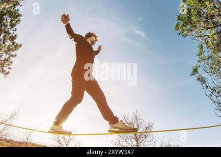 Un homme, vieilli avec une barbe et portant des lunettes de soleil, se balance sur une slackline en plein air entre deux arbres au coucher du soleil sur fond bleu ciel Banque D'Images