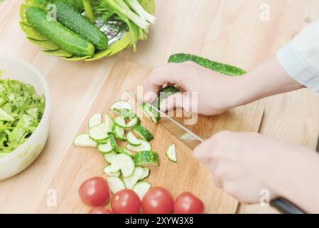 Gros plan des mains femelles coupées en concombres frais coupés sur une planche à découper en bois à côté des tomates roses. Le concept de cuisine végétarienne maison et Banque D'Images