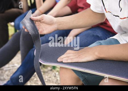 Un jeune garçon à genoux colle le griptape sur un skateboard en compagnie d'amis par temps ensoleillé. Préparation d'une planche à roulettes pour une compétition de skatepark Banque D'Images