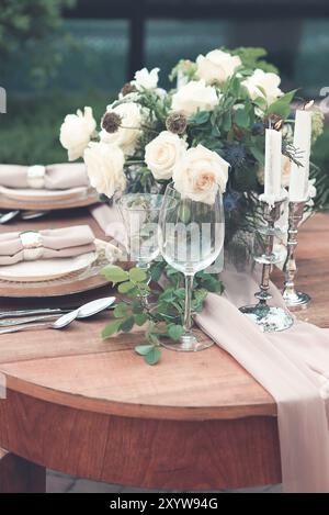 Bougies et vase avec des roses blanches placées sur la table ronde près des verres à vin pendant la date romantique dans le jardin Banque D'Images