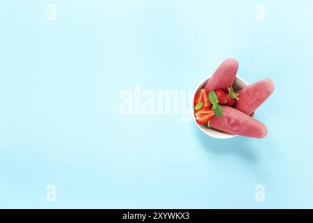 Vue de dessus sur bol avec sorbet glacé rose fruité crème glacée sur bâton de plastique avec fraise, menthe sur fond bleu. Fruits mélangés congelés avec yaourt i Banque D'Images