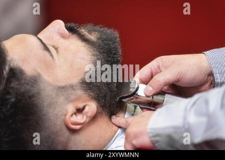 Barbier rasant la barbe d'un bel homme barbu avec un rasoir électrique au salon de coiffure Banque D'Images