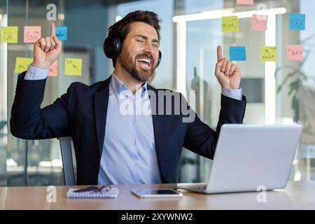 Heureux homme d'affaires appréciant la musique sur les écouteurs au bureau, entouré de notes autocollantes. Multitâche professionnel et détente pendant les heures de travail. Énergie positive et productivité dans le travail moderne Banque D'Images
