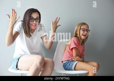 Heureuse mère et sa petite fille assise sur les chaises par le mur gris. Portrait de Mère et fille avec des lunettes Banque D'Images